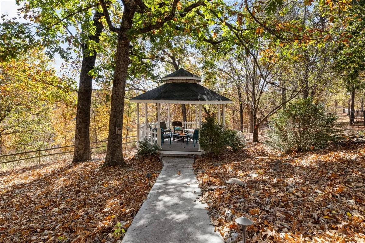 A tranquil exterior scene featuring a gazebo nestled among mature trees with fallen autumn leaves carpeting the ground. The setting offers a peaceful spot for relaxing, reading, or enjoying nature, and highlights the changing seasons with its colorful foliage.