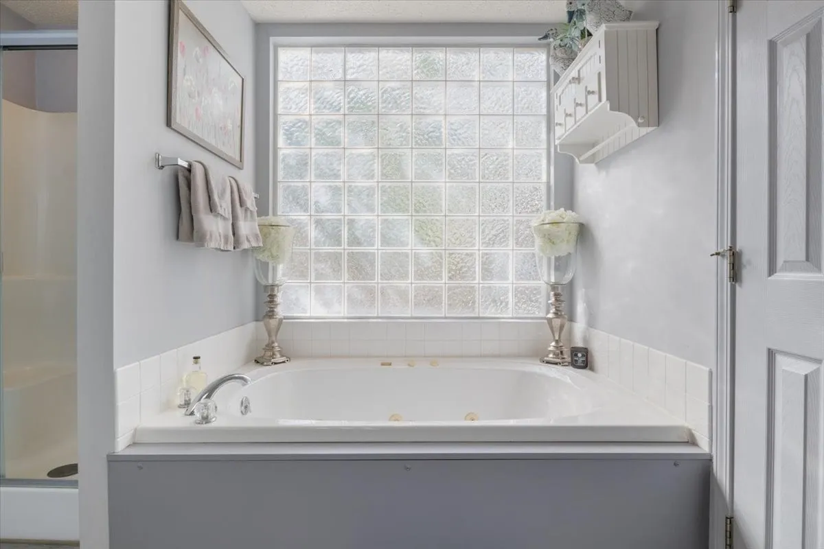 A spa-inspired bathroom with a jacuzzi tub set beneath a glass block window, complemented by decorative lamps. The inviting setup promises a serene, spa-like bathing experience in a private, well-lit environment.