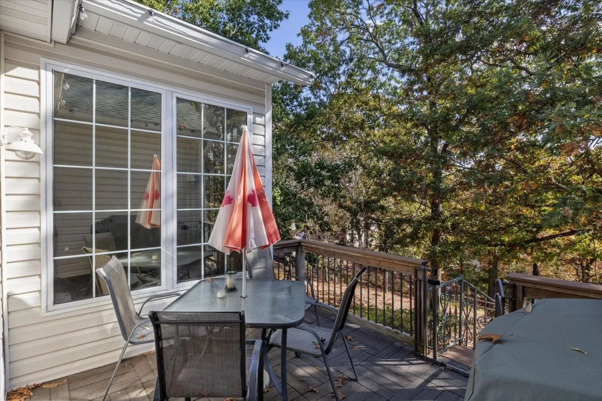 A serene outdoor deck with a glass dining table, red and white umbrella, and wooded surroundings. The space is perfect for outdoor dining, socializing, or enjoying a sunset among the trees.