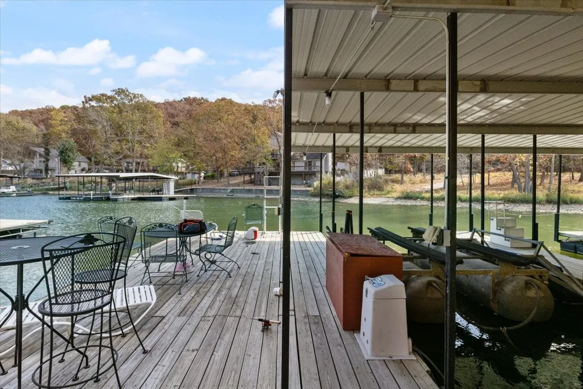 A picturesque dock with a boathouse extends over the tranquil water of Lake of the Ozarks, surrounded by vibrant autumn foliage. The dock features a comfortable seating area perfect for relaxing, while the lake offers opportunities for fishing, boating, and soaking in sunset views.
