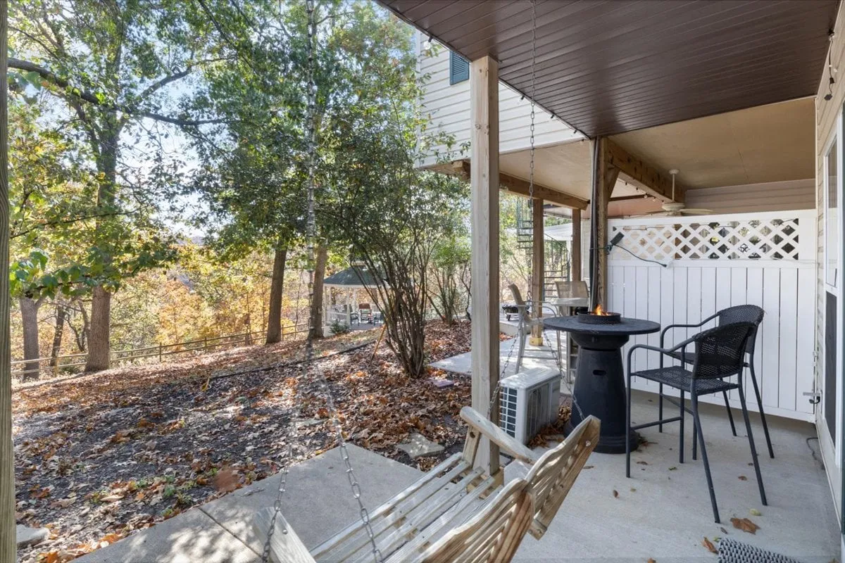 An inviting exterior scene showing a swinging bench, black chairs, and a fire pit nestled amongst surrounding trees. The arrangement suggests a space designed for evening gatherings, fireside chats, and quiet reflection in nature.