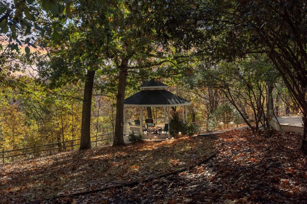 A peaceful gazebo nestled among mature trees with fallen autumn leaves. The gazebo offers a cozy retreat for relaxing, reading, or enjoying the sunset, surrounded by the beauty of nature.