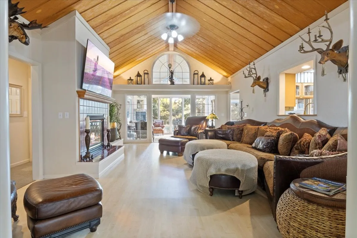 A soaring living room with a high vaulted wooden ceiling, a large sectional couch perfect for group seating, a roaring fireplace, a TV for entertainment, and expansive windows leading to an outdoor deck. The setup is ideal for both lively gatherings and relaxed family moments.